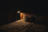 Bedside lamp casting a warm glow over an unmade bed in a dark room