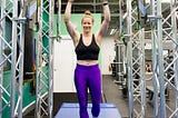 The author stands amidst a bunch of american ninja warrior obstacles in black tank top and purple pants, smiling at the camera