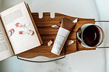 Photo of a bathtub with a wooden bath tray. On the bath tray is an open book, rose petals, a copper bottle of face mask mud, and a red glass of wine.