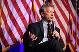 Senator Rand Paul sitting down with a microphone in hand with an American flag behind him.