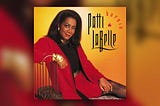 Patti LaBelle’s ‘Burnin’’ album cover: she sits on a chaise lounge with a backing made of thin, curved metal railing. She wears a red skirt suit, with a black top, gold bangles on her right wrist, a gold geometric necklace, and a shiny, gold belt.