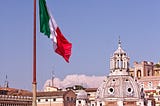 A green, white, and red flag is flying above an ancient stone building with an ornate domed top, surrounded by shorter stone buildings.