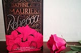 image shows a book — Rebecca by Daphne du Maurier, standing on its end beside a red rose.