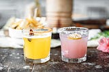 An orange cocktail with a chilli flake rim and a dried chilli garnish, sits next to a pink cocktail with a salt rim and a dried lemon slice garnish.