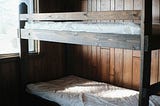A photo of a wooden bunk bed with bare mattresses and a snipped of window.