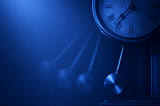 Monochromatic blue illustration of a vintage pendulum clock with Roman numerals. The pendulum appears in multiple blurred positions, creating the impression of continuous motion. Soft blue light fades across the left side of the image, leaving open space and emphasizing themes of shifting cycles, changing priorities, the passage of time, and institutions reacting to forces already in motion.