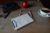photograph of wooden table with red candle, clipboard with pen drawing, two dice, one pen, one coffee (cappuccino, chocolated on the closest side) and one copy of the Monster Manual