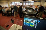 A laptop screen on top of the lectern at the firearms instructors conference behind which there are the attendees sitting at tables (this part of the picutre is blurred). The laptop is showing a slide of a presentation with the text ‘Firearms Instructors Conference — Guardians of the Standard’, the slide has the picture of a group of firearms instructors in the background, set outside in a teaching setting.