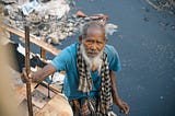 An old man next to a polluted river looks at the camera while holding his cane.