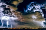 Photo of a storm darkened sky with mixtures of Dark blue, brown, black, and yellow coloring the stormy sky, while bolts of lightning erupt from storm clouds to touch the darkened earth of the city.