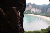 A man in a climbing harness looks down on a Thai bay