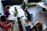 Five women sitting at a large table with laptops, taking notes in an open office space