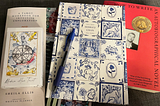 A Tarot card deck, a Tarot workbook, a blue pen on top of a planner, and a copy of Alexander Chee’s essay collection, How to Write an Autobiographical Novel. The card deck is named “Modern Hue” and features watercolor illustrations.