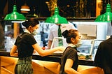 Picture of servers wearing masks picking up food at a counter with green swinging lights overhead, Picture