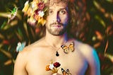 A shirtless, bearded man stands with flowers, butterflies and hummingbirds