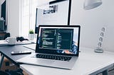 A MacBook with lines of code on its screen on a busy desk.