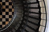 View from above of a black spiral staircase against a white wall, and a black and white checked tile floor