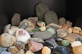 a collection of rocks of different shapes, colors, and sizes, with a tiny bluebird figurine and a trinket of a dragon hatching from an egg.