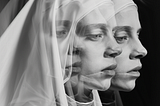 A black-and-white double-exposure portrait of a woman in a white veiled hood and robe, shown twice in overlapping profile — one image slightly translucent and layered over the other, creating a ghostly, fractured effect. Her eyes are downcast in both versions, with soft, ethereal lighting against a dark background, evoking a sense of duality or a self splitting apart.