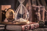 A centuries old hourglass sits atop an old leather-bound book on a desk.