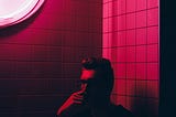 man sits in a corner, walls of ceramic tile, a large white mirror sits over his head almost out of view, the lighting is a bleak, dim red and almost everything, including the man, is darkly shadowed