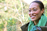 Woman in green scarf smiles off the camera with a background of trees.