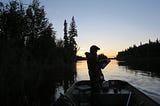 A person stands in a boat in silhouette with forest and water and the beginning of a sunrise in the background.