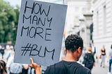 A Black man at a protest faces away from the camera holding a hand-written sign that says “How Many More #BLM”