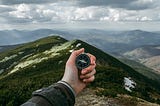 Hand holding a compass in front of a backdrop of green mountains stretching into the distance.