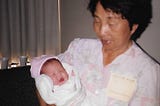 A young grandma lovingly cradles her newborn granddaughter swaddled in blankets and a pink beanie.
