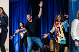 A group of people at a conference. On the left, five people hold blue dolls. In the center, two individuals pose energetically on stage. On the right, five people stand in front of a branded backdrop, smiling.