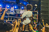 Uma criança negra pequena, apoiada nos ombros do pai, em frente a um trio elétrico durante o carnaval de Salvador. As pessoas em volta olham e sorriem para ela.