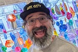 Closeup of the author with a big open-mouth smile. He’s wearing a hat, has a long graying beard and glasses, and is standing in front of colorful glass art.