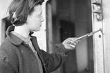 A black and white image. A woman stands in a Tyneside shipyard, her upper-body is visible and she is holding her arm out, holding a tool for catching rivets. She is wearing a coat and overalls with a headscarf and looks towards the work she is doing