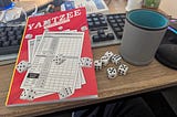 An off-brand Yahtzee scorepad (the “H” is covered by a rolling die, to avoid trademark offense), six dice and a leatherette dice cup sitting on a desk in front of a computer keyboard.