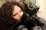 A woman with long, curly black hair wearing a black leather jacket (Karen Dendy Smith) and holding a black cat with yellow eyes (Hockey Puck).