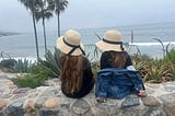 Two young girls sit on a stone wall overlooking the Pacific ocean.