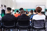 The backs of three students with different skin tones seated side by side in the back of a classroom full of students facing a teacher and a white board.