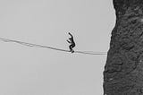 Looking up from below to see a man in the distance walking on a tightrope from the edge of a cliff.