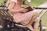 A young girl sitting on sn old bench reading a book.