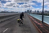 A photo of a young man on a bicycle, riding along a long highway, with a blue sky dotted with clouds above, and a city in the distance ahead