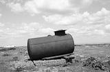 A black and white photo of an abandoned cylindrical tank in a countryside.