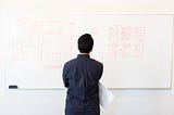 Man with his back facing us stands thinking in front of a whiteboard with writing on it