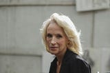 A woman with white hair and wrinkles, looking directly at the camera with a pensive look on her face, against a cement block wall.