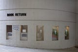 An exterior curving light tan concrete structure with a “Book Return” sign at the left side. Underneath the sign, there are two book returns. To the the right, there are three small modern glass panels offering a peek into the library.