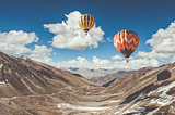 Two hot air balloons in the clear blue sky with big white clouds. A meadow and mountains below.