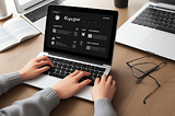 Image of a laptop on a desk with three hands typing at the keyboard. Glasses with one eye glass are next to it.