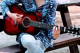 A red guitar. The woman playing the guitar is sitting on a picnic bench and her face is not in the frame