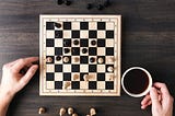 A chess game on a small board framed by two hands and a cup of coffee.