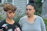 teen with one badly bruised eye speaks into a reporter’s microphone. his mom stands next to him. they are in front of a house with wooden siding and flowering bushes.